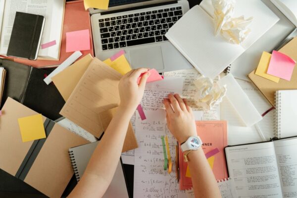 Chaos im Kopf kann durch einen Schreibtisch im Chaos begünstigt werden: Zwei Hände kleben Post-Its auf Zettel, es liegen viele (zerknüllte) Zettel, Notizbücher und Ringbücher herum, Laptoptastatur schaut heraus